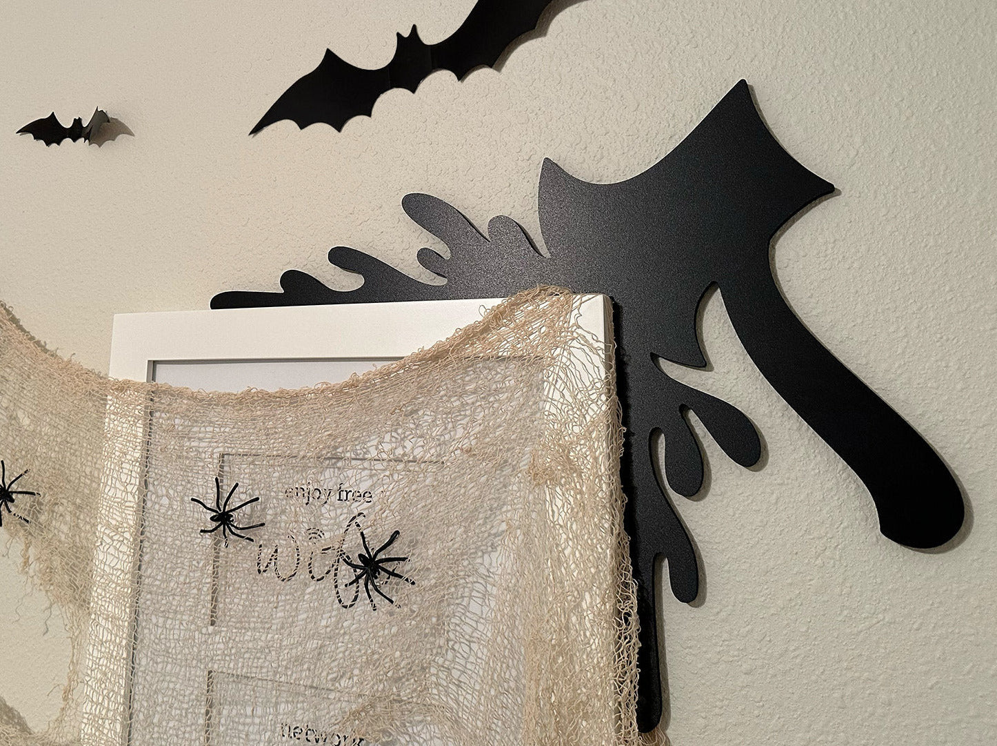 A black door frame topper, designed with the silhouette of an axe and blood splashes, is seen on top of a black frame. The frame is covered in spider webs. The wall behind is beige.