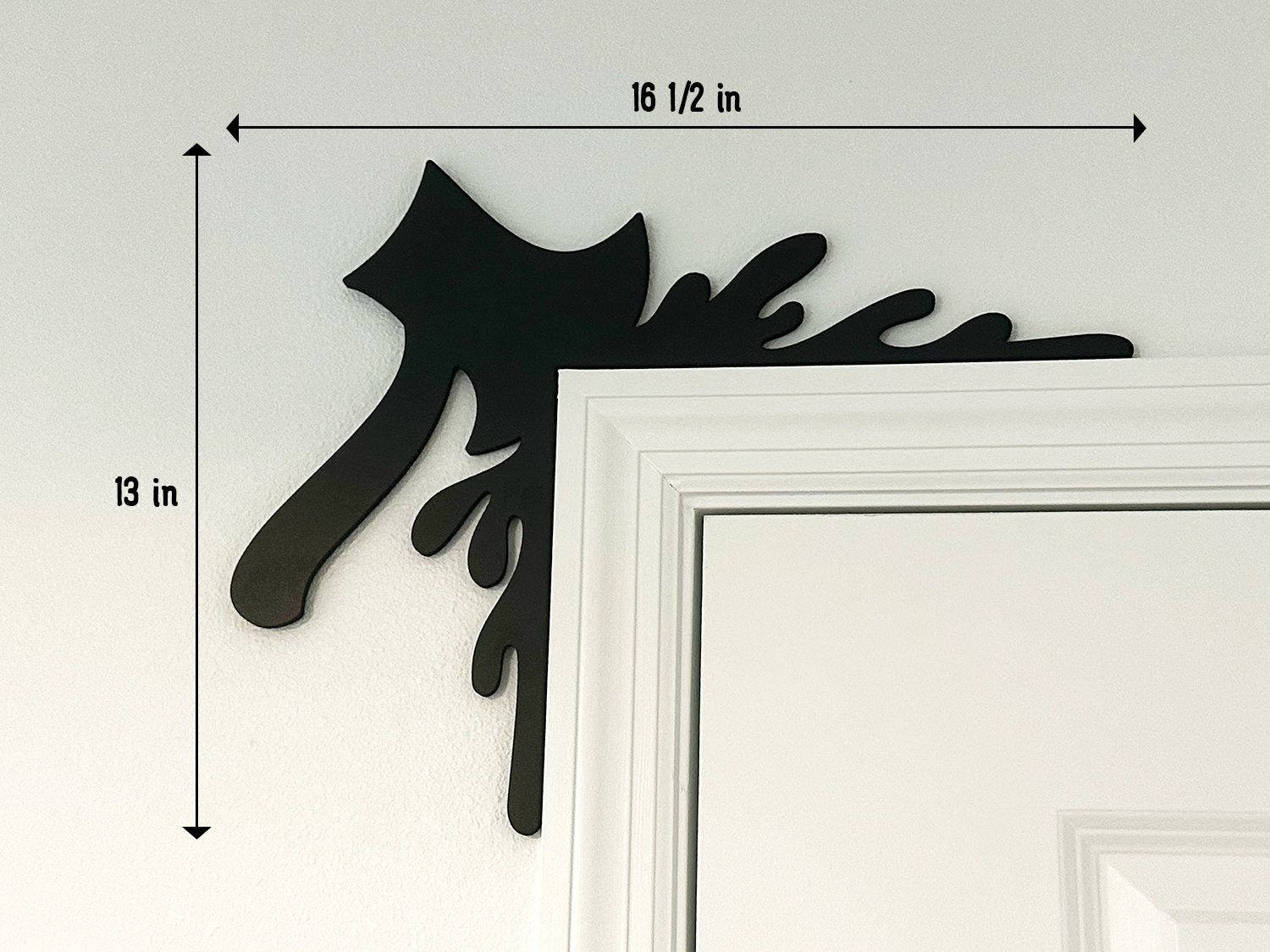 A black door frame topper, designed with the silhouette of an axe and blood splashes, is seen on top of a black frame. The frame is covered in spider webs. The wall behind is beige.