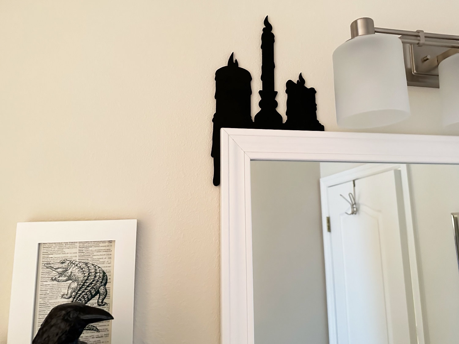 A black door frame topper, designed as a silhouette of three dripping wax candles, is seen on top of a white door frame. Spooky bats and spiders surround the topper. The wall behind is beige.