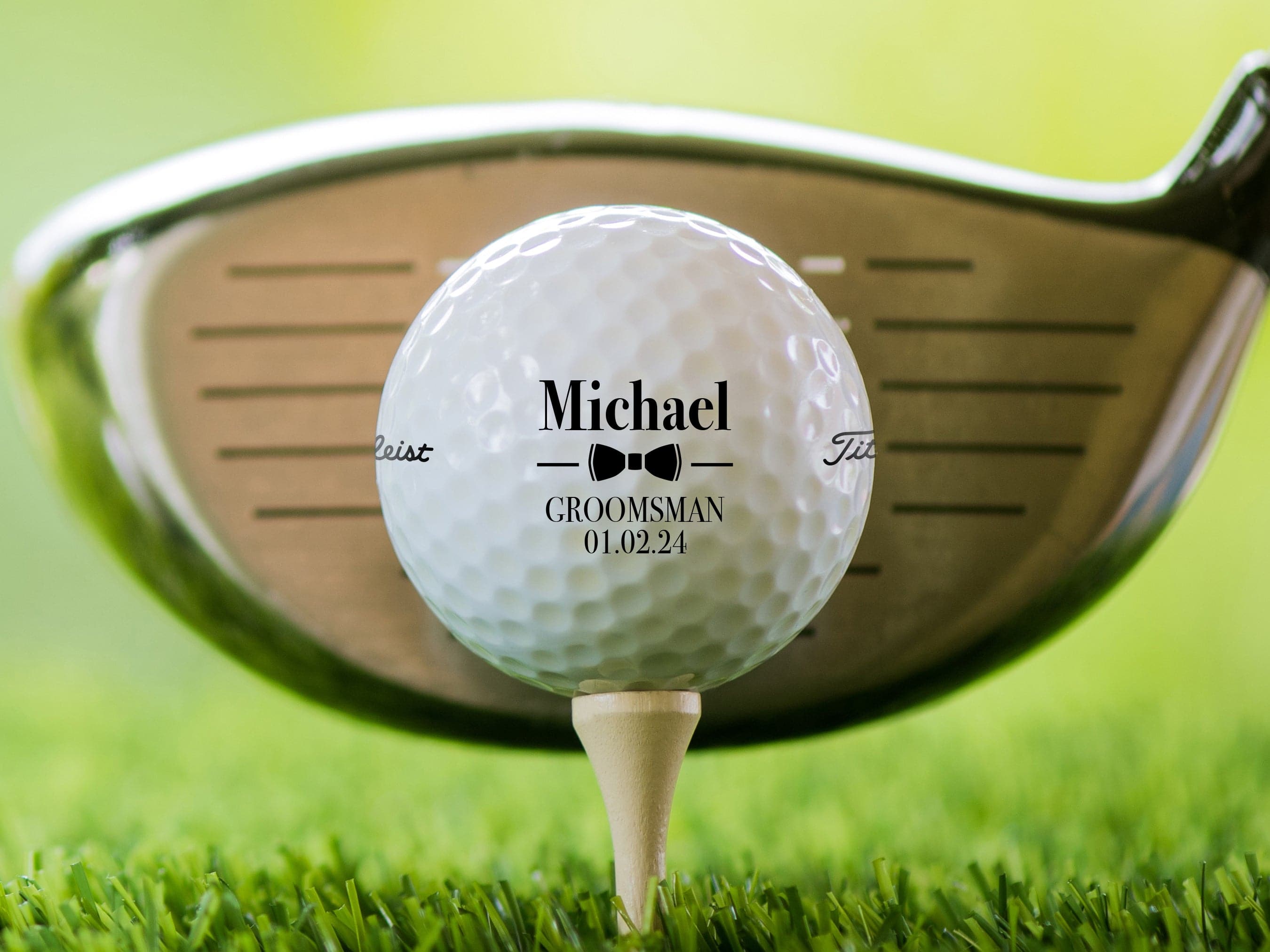 single white titleist golf ball with customizable personalized groomsmen bowtie design on white golf tee on golf course grass background