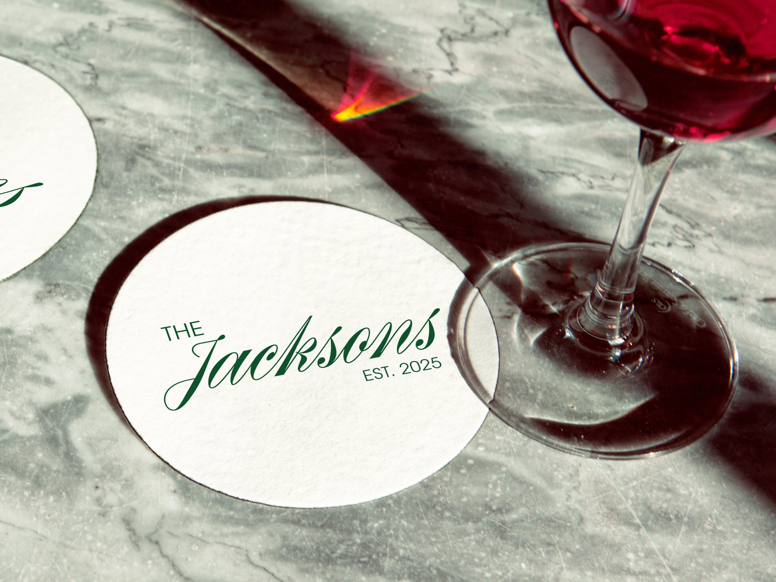 A stack of coasters are shown on a white background. Coasters feature Formal Last Name and Established Date design. This design has a white background and the happy couples last name and wedding year in emerald green writing.