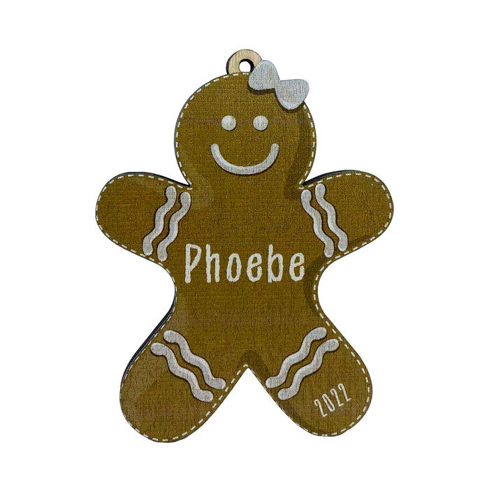 Ornament shown with Gingerbread Girl design. The ornament is wooden and shaped like a gingerbread boy. Name and year are written on ornament in white.