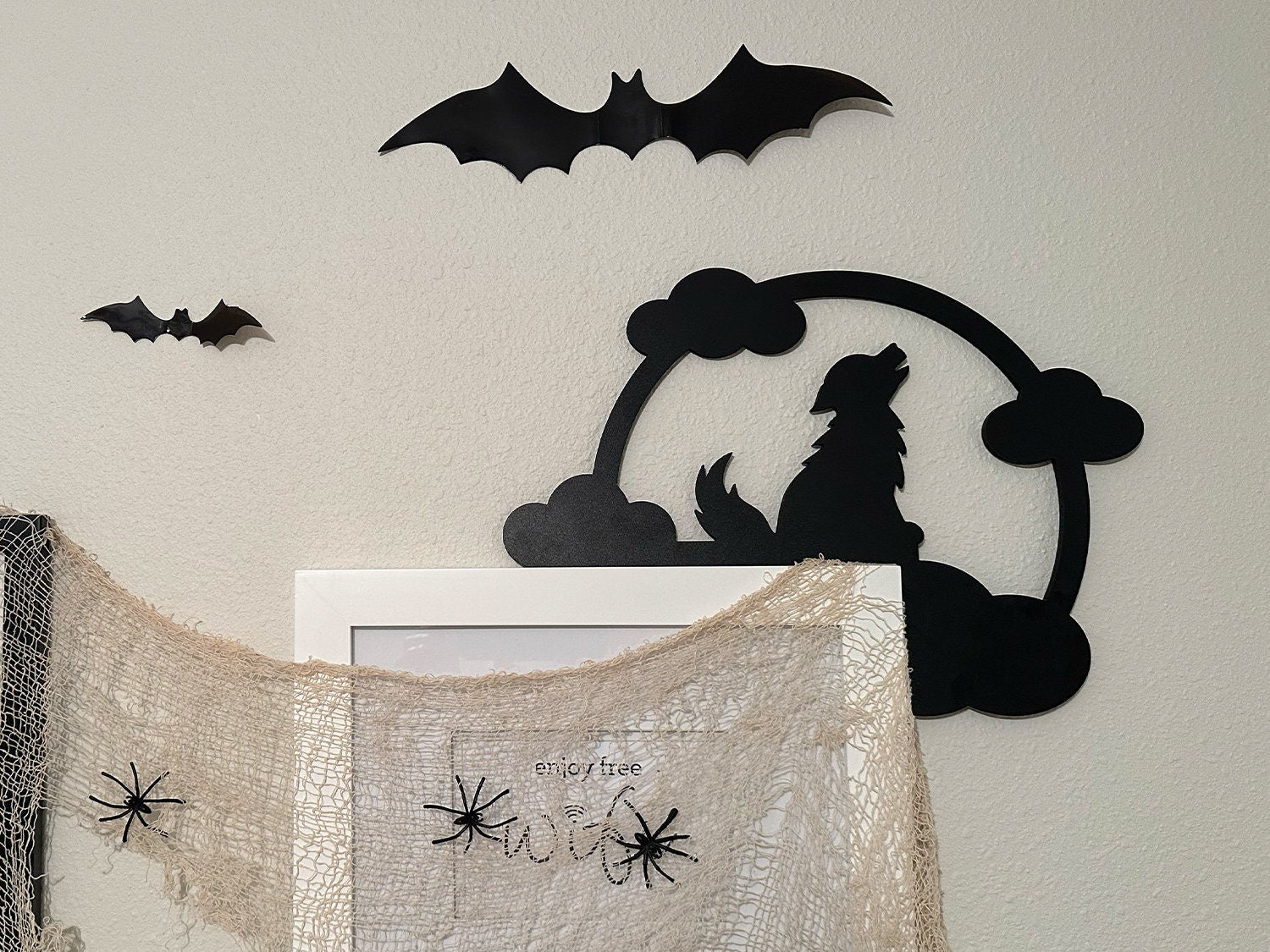 A black door frame topper, designed with the silhouette of a howling wolf, full moon, and clouds, is seen on top of a white door frame. The frame is covered in black spider webs and fake spiders. The beige wall behind has fake bats attached to it.