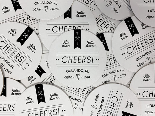 Multiple coasters spread in all different directions. Coasters feature CHEERS! Modern Wedding design. Coasters show married couple names and first name initials, the word “CHEERS!”, and the wedding location and date. Design is printed in black on a white coaster. These coasters also have decorative keys, lines, and dots on it.