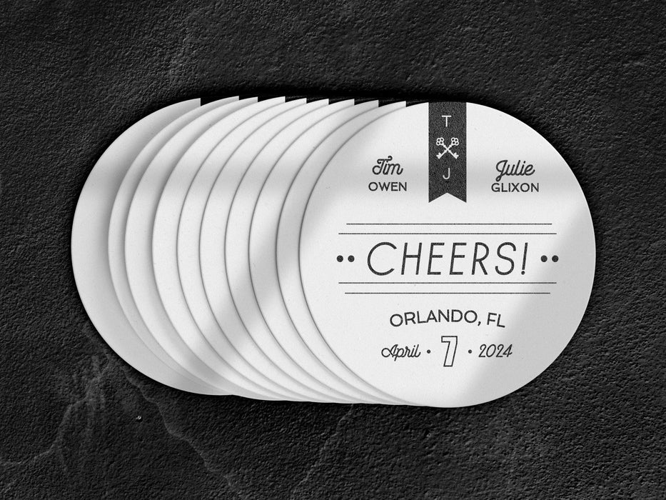Stack of coasters spread out on black marble background. Coasters feature CHEERS! Modern Wedding design. Coasters show married couple names and first name initials, the word “CHEERS!”, and the wedding location and date. Design is printed in black on a white coaster. These coasters also have decorative keys, lines, and dots on it.