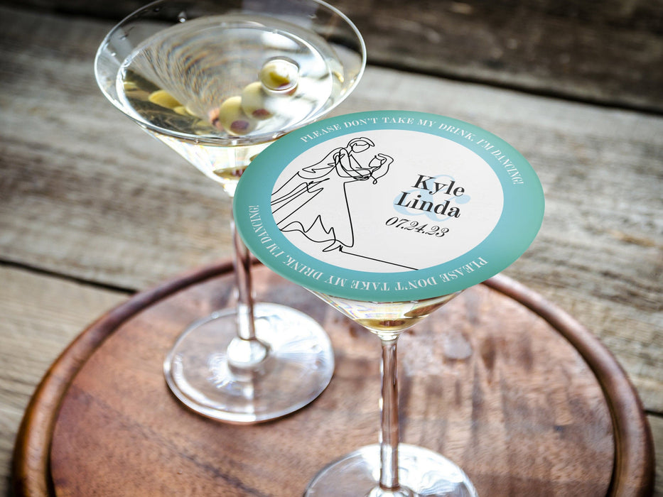 Don't Take My Drink coaster placed on top of martini glass on round wooden tray. Coasters say Please Don't Take My Drink, I'm Dancing in white around a teal border with an illustration of a married couple dancing beside their names and wedding date.