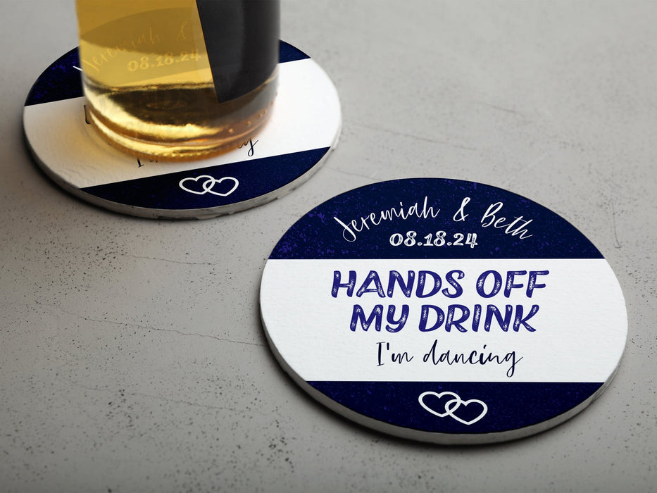 One coaster has a drink on it and an empty coaster sits beside it. Coasters say married couple names, wedding date, HANDS OFF MY DRINK I'm dancing. Coaster design is printed in purple and black on a white coaster.