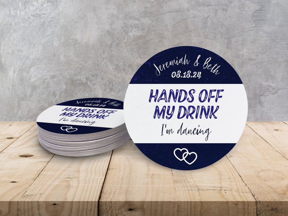 A stack of coasters by a single coaster on wooden table. Coasters say married couple names, wedding date, HANDS OFF MY DRINK I'm dancing. Coaster design is printed in purple and black on a white coaster.