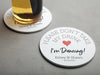 One coaster has a drink on it and an empty coaster sits beside it. Coasters say please don't take my drink I'm dancing with married couple names and wedding date below. Red heart featured in the middle of coaster.