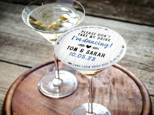 Don't Take My Drink coaster placed on top of martini glass on round wooden tray. Coasters say Please Don't Take My Drink I'm Dancing with wedding couple names and wedding date. The words, place on top of your drink while away, act as a border around coaster.