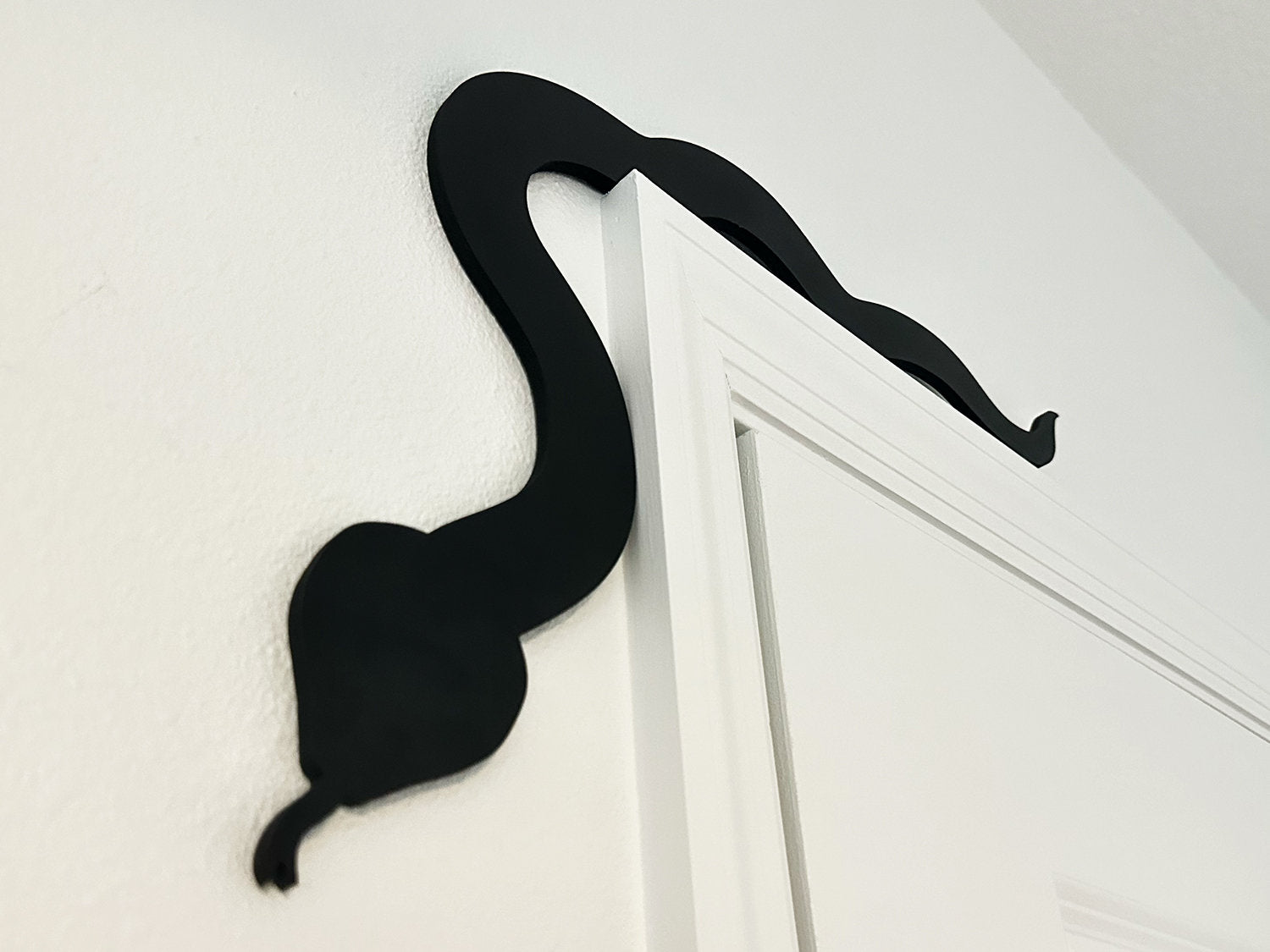 A black door frame topper, designed with the silhouette of a slithering snake, is seen on top of a white door frame decorated with fake spiders and spider webs. The wall behind is beige with fake bats on it.