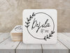 A stack of coasters by a single coaster on a white wooden table. Coasters feature Floral Family Name design. Design has a simple, circular, floral frame around the words "DeJulio Est. 2022". Design is printed on a white square coaster.