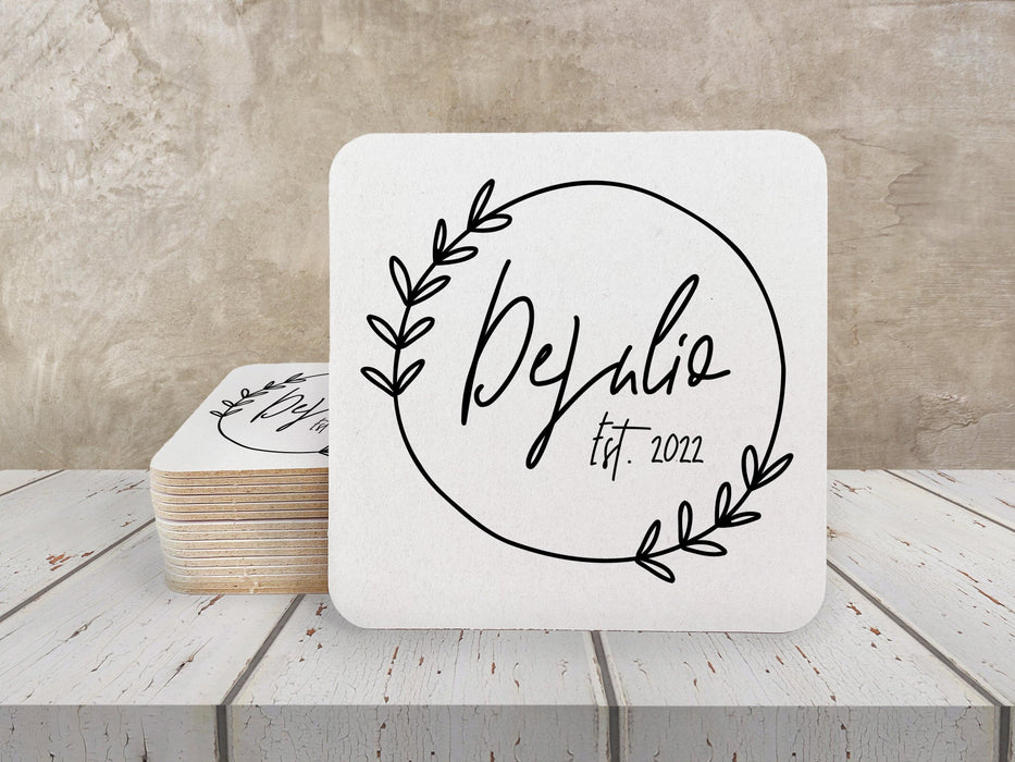 A stack of coasters by a single coaster on a white wooden table. Coasters feature Floral Family Name design. Design has a simple, circular, floral frame around the words "DeJulio Est. 2022". Design is printed on a white square coaster.