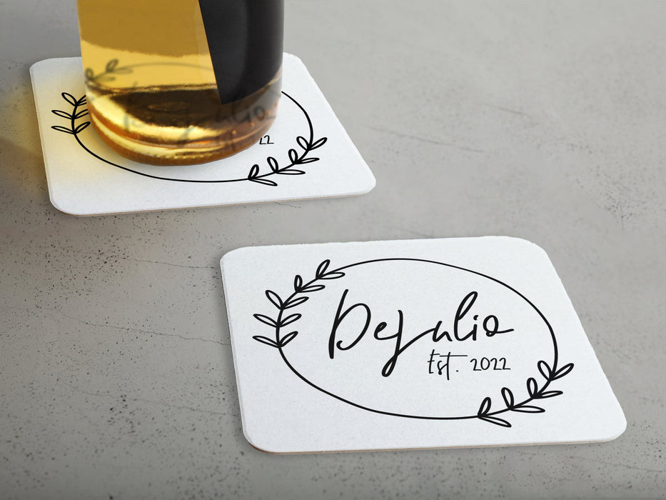 One coaster has a drink on it and an empty coaster sits beside it. Coasters feature Floral Family Name design. Design has a simple, circular, floral frame around the words "DeJulio Est. 2022". Design is printed on a white square coaster.
