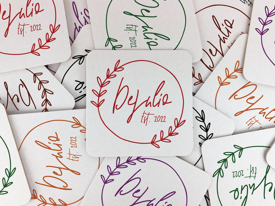 Multiple coasters shown in the following colors: Black, Brown, Green, Orange, Purple, and Red. Coasters feature Floral Family Name design. Design has a simple, circular, floral frame around the words "DeJulio Est. 2022". Design is printed on a white square coaster.