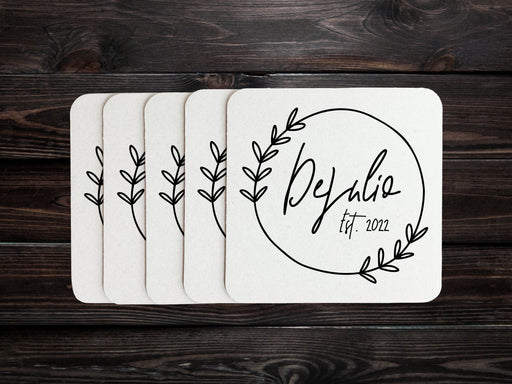 A stack of coasters are spread out on a dark wooden table. Coasters feature Floral Family Name design. Design has a simple, circular, floral frame around the words "DeJulio Est. 2022". Design is printed on a white square coaster.