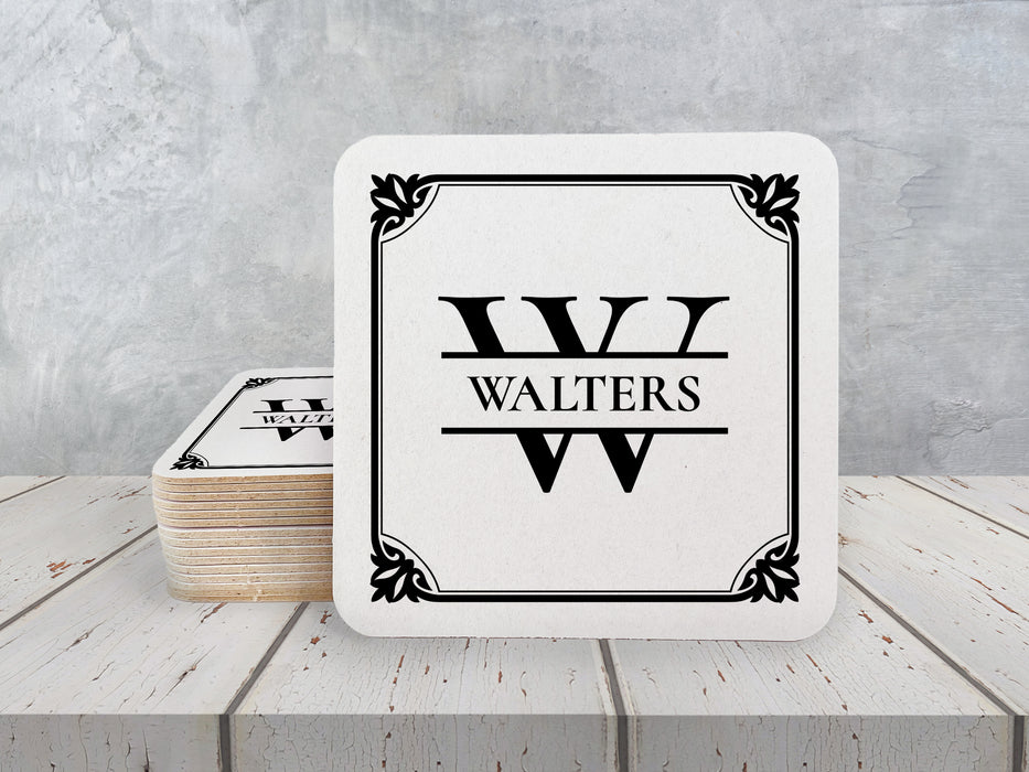 A stack of coasters by a single coaster on a white wooden table. Coasters feature Framed Monogram Family Name design. Design has a square ornamental frame around a monogram for the last name "Walters". Design is printed on a white coaster.