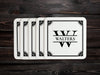 A stack of coasters are spread out on a dark wooden table. Coasters feature Framed Monogram Family Name design. Design has a square ornamental frame around a monogram for the last name "Walters". Design is printed on a white coaster.