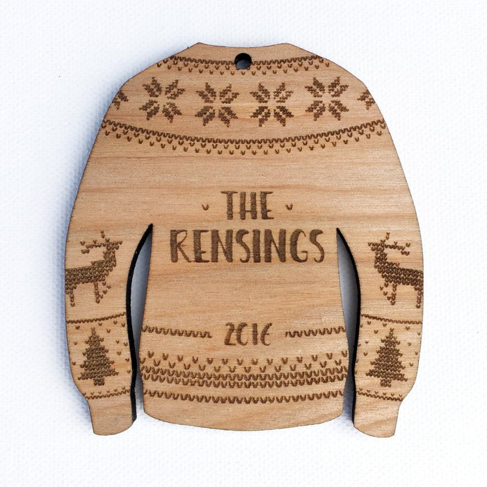 Ornament features an Ugly Sweater design. Design is made in a sweater shape with laser etched stitched decorative details. Decorative reindeer and Christmas trees are on sleeves. Custom last name and year are laser etched in the center.