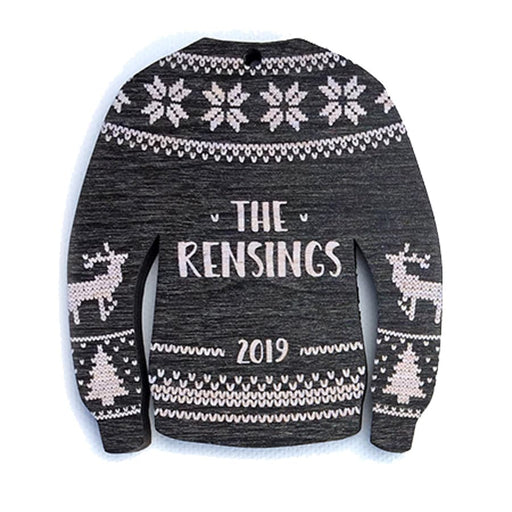 Ornament features an Ugly Sweater design. Design is black and made in a sweater shape with white stitched decorative details. Decorative reindeer and Christmas trees are on sleeves. Custom last name and year are printed in white in the center.