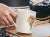 woman's hand holding a 15 ounce ceramic mug featuring artwork of a bouquet of pastel spring flowers on a kitchen table pouring a coffee pot