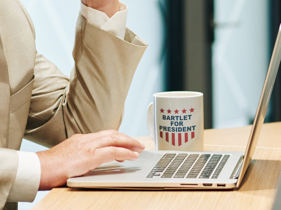 businessman sitting at desk with computer with a white mug with red white and blue American design with typography that says Bartlet for president with Stars and Stripes