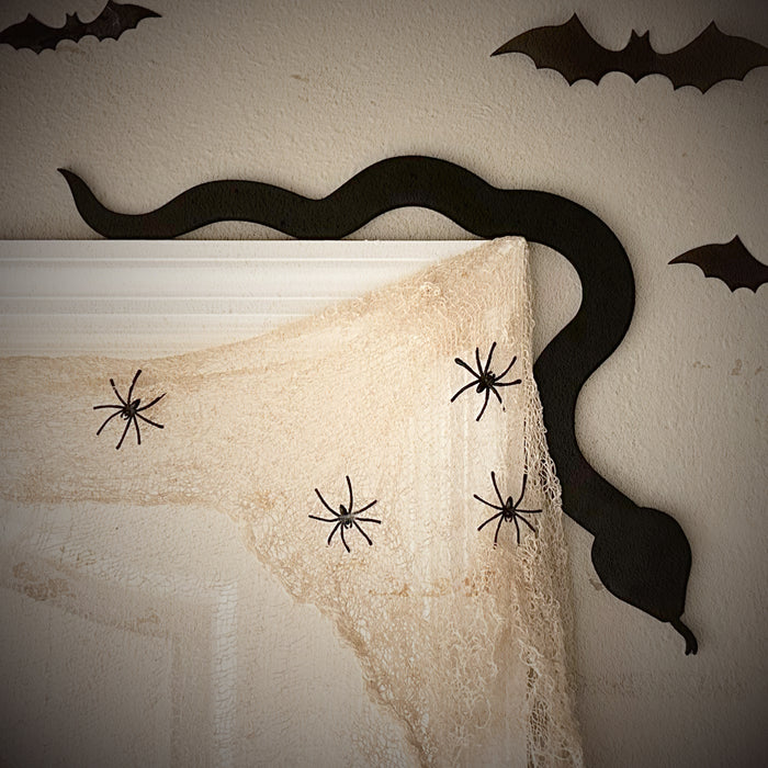 A black door frame topper, designed with the silhouette of a slithering snake, is seen on top of a white door frame decorated with fake spiders and spider webs. The wall behind is beige with fake bats on it.