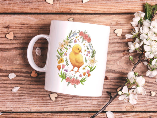 15 ounce white ceramic mug with spring easter art of a baby chick surrounded by various colorful flowers sitting on a wooden table next to a bunch of white flowers, and heart shaped petals