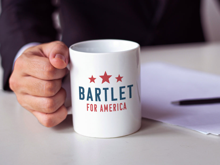 Business man holding white mug with red white and blue American design with typography that says Bartlet for America with Red Stars on white table with paperwork and pen