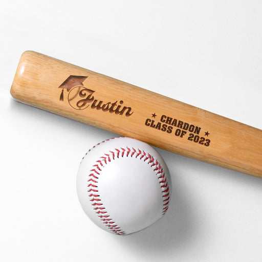 wooden mini baseball bat with custom laser engraved design that features a name with a graduate cap and says "Justin, Chardon, Class of 2023" on a white surface next to a baseball