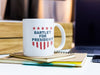 white mug with red white and blue American design with typography that says Bartlet for president with Stars and Stripes sitting ontop of notebooks at desk with computer and phone