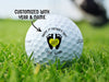 Single white titleist golf ball with first Father's Day footprint design in yellow variation on a grassy golf course. The text "customize with year and name" is above the ball with an arrow pointing toward it.