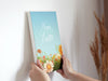 two hands holding a 8x10 canvas of happy easter artwork of a spring meadow against a white living room wall with a house plant in the background