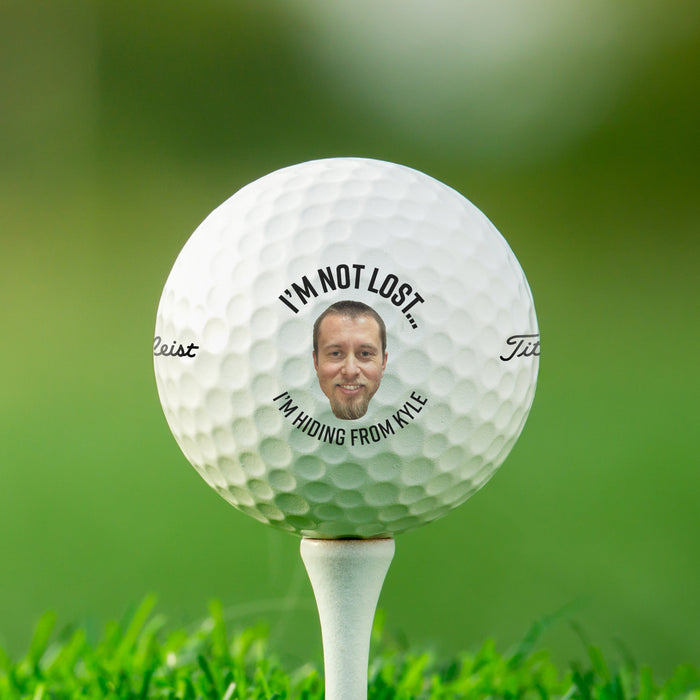Single white Titleist golf ball with I'm Not Lost, I'm Hiding From design on white golf tee with golf course grass in the background.