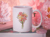 15 ounce ceramic mug featuring artwork of a bouquet of pastel spring flowers on a book surrounded by pink items such as a wallet, a pendant, and pink flowers