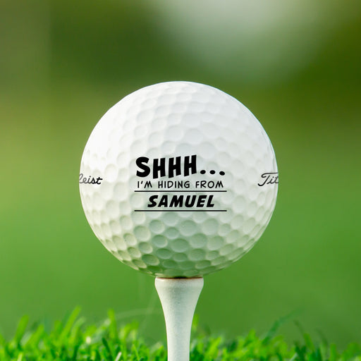 Single white Titleist golf ball with Shhh I'm Hiding From design on white golf tee with golf course grass in the background