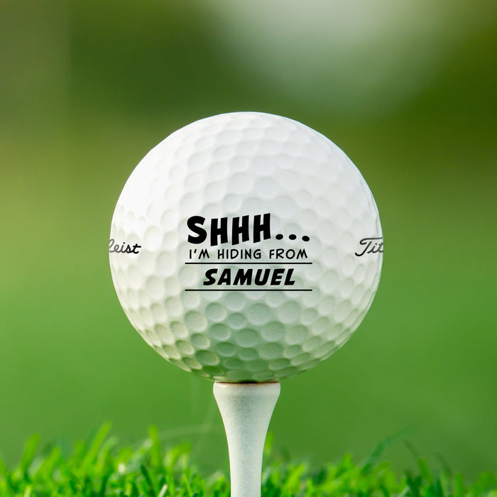Single white Titleist golf ball with Shhh I'm Hiding From design on white golf tee with golf course grass in the background