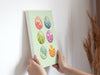 two hands holding a 8x10 canvas with colorful easter artwork of rows of eggs and a baby chick against a white wall with a house plant in the background