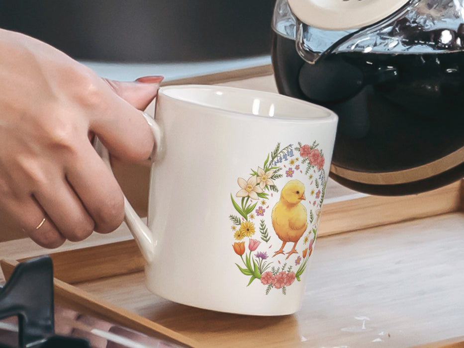 woman's hand holding a 15 ounce white ceramic mug with spring easter art of a baby chick surrounded by various colorful flowers in a kitchen pouring a coffee pot into the mug