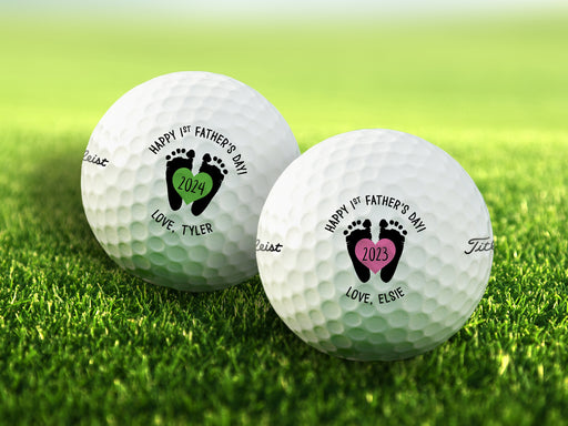 Two white Titleist golf balls with first Father's Day footprint designs on top of golf course grass background