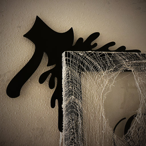 A black door frame topper, designed with the silhouette of an axe and blood splashes, is seen on top of a black frame. The frame is covered in spider webs. The wall behind is beige.