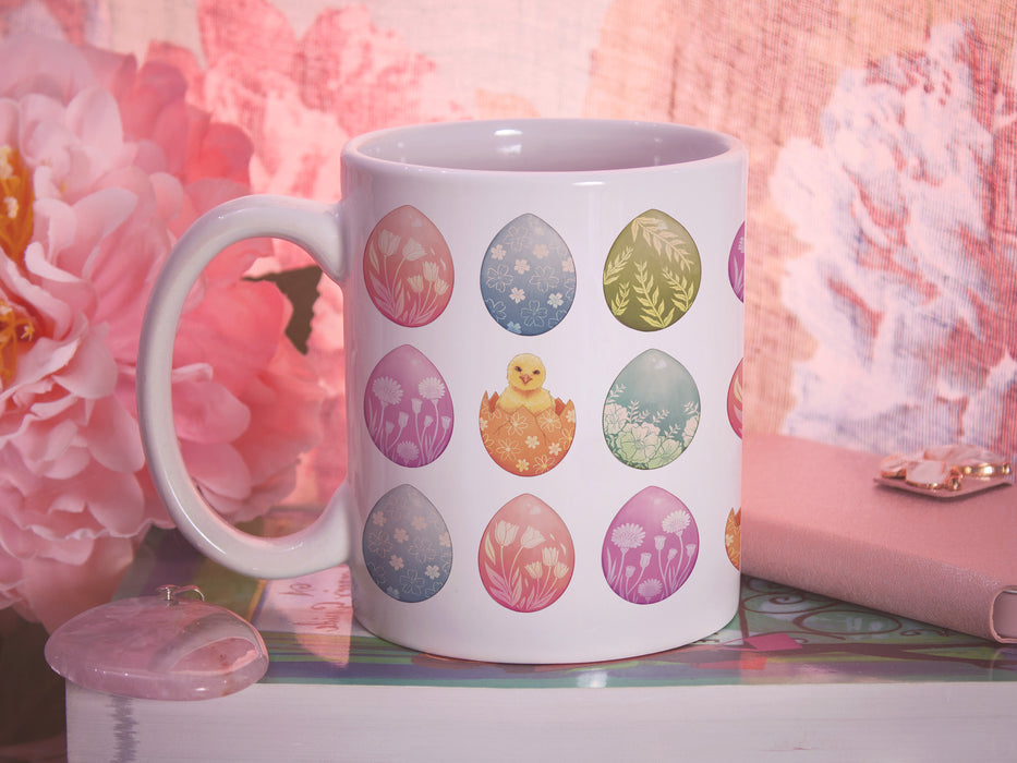 15 oz white ceramic mug with an easter pattern of floral decorated eggs with a baby chick popping out of an egg on a book surrounded by pink items such as a wallet, a pendant, and a flower