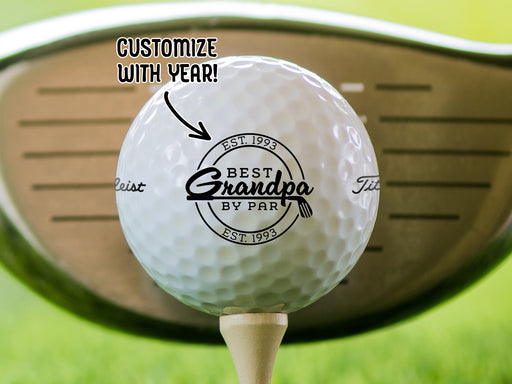 Single white titleist golf ball with Best Grandpa By Par design on wooden golf tee with golf club and golf course grass in the background. The text "customize with year" is above the ball with an arrow pointing toward it.