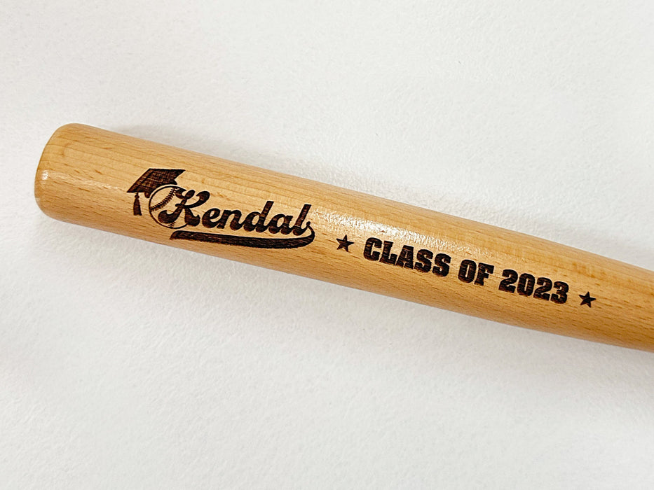 wooden mini baseball bat with custom laser engraved design that features a name with a graduate cap and says "Kendal, Class of 2023" on a white surface