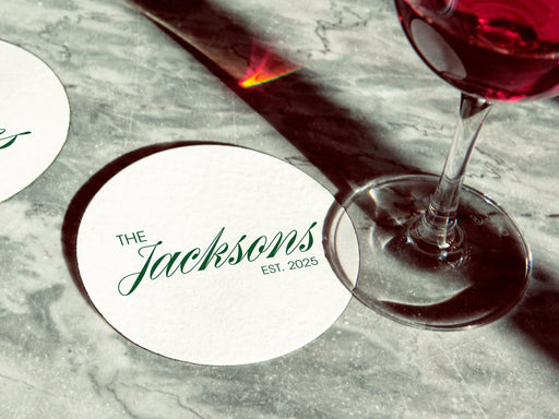 Two coasters are shown with a wine glass on top of one on a marble table. Coasters feature Formal Last Name and Established Date design. This design has a white background and the happy couples last name and wedding year in emerald green writing.