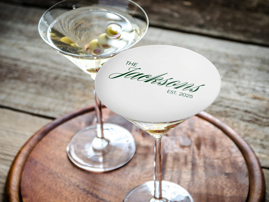 One coaster is shown being used as drink cover on top of a martini glass. Coaster features Formal Last Name and Established Date design. This design has a white background and the happy couples last name and wedding year in emerald green writing.