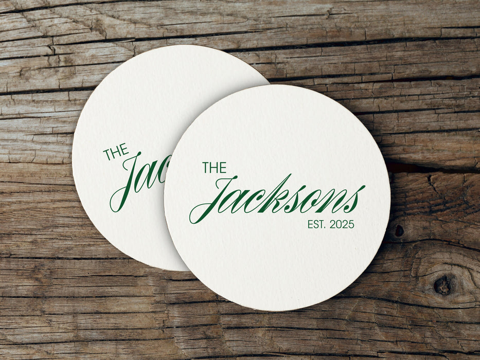 Two coasters are shown stacked on top of each other on wood table. Coasters feature Formal Last Name and Established Date design. This design has a white background and the happy couples last name and wedding year in emerald green writing.