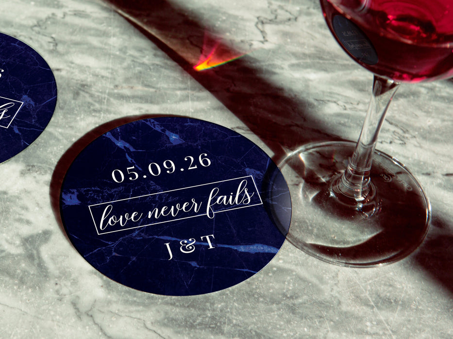 Two coasters are shown with a wine glass on top of one on a gray marble table. Coasters feature Love Never Fails design. This design has a dark blue marble background and the happy couples first name initials and wedding date in white writing.