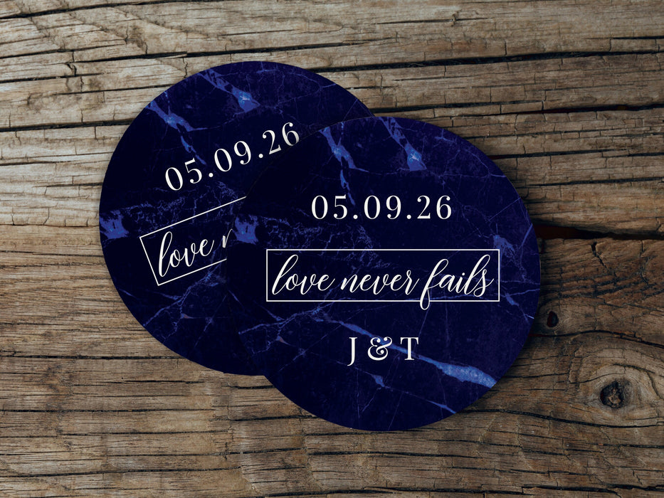 Two coasters are shown stacked on top of each other on wood table. Coasters feature Love Never Fails design. This design has a dark blue marble background and the happy couples first name initials and wedding date in white writing.
