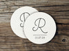 Two coasters are shown stacked on top of each other on wood table. Coasters feature Monogrammed Initial design. This design can be customized with a large initial and date in black writing on a plain, white background.